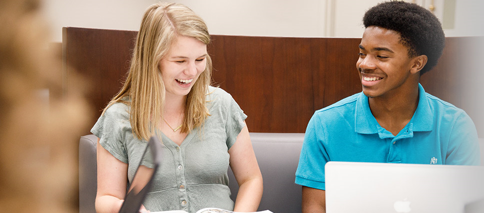 Two students in a library laugh as they study with on laptops