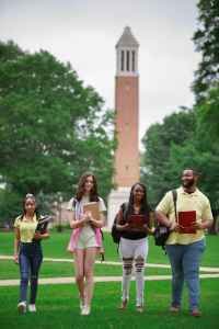 Four smiling high school students, with books in hand, walk across the quad to their next college class, as Denny Chimes is visible behind them.