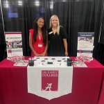 An Early College ambassador poses with an Early College recruiter, at a college fair. They smile while standing behind a red table, covered with UA Early College banners, shakers, pins, highlighters and brochures.