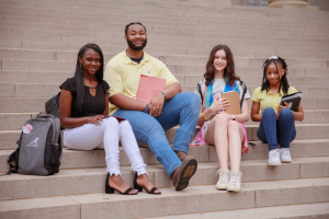 Four high school students sit on the steps of a library, on The University of Alabama campus, and catch up with each other, between their college courses.